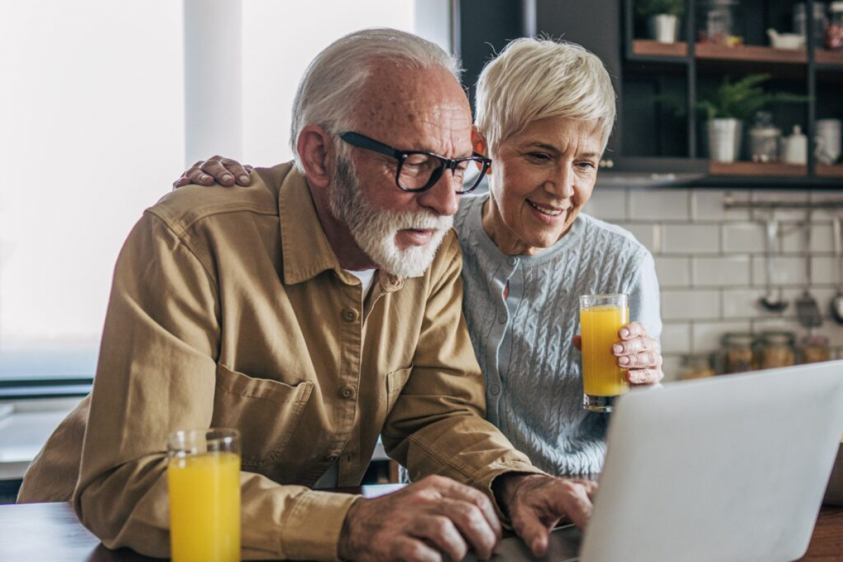 The senior man and his wife are standing side by side in the kitchen, enjoying a moment together. Both are looking at the laptop screen, sharing the experience. Beside them is a glass of fresh orange juice, which they’re both savoring.